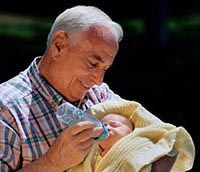 Fotografía de un abuelo cargando a su nieto recién nacido, y alimentándolo por medio de un biberón